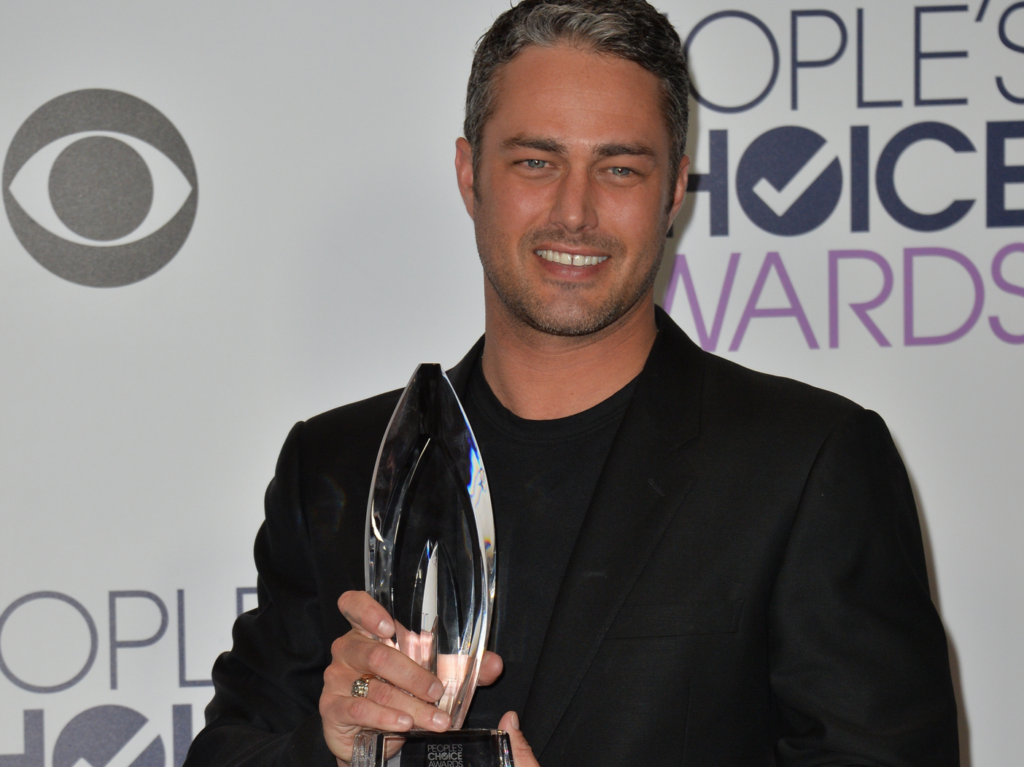 Taylor Kinney poses with an award in a black suit with black shirt