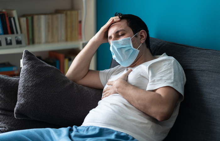 A sick man wearing a face mask holds his chest while sitting on a couch