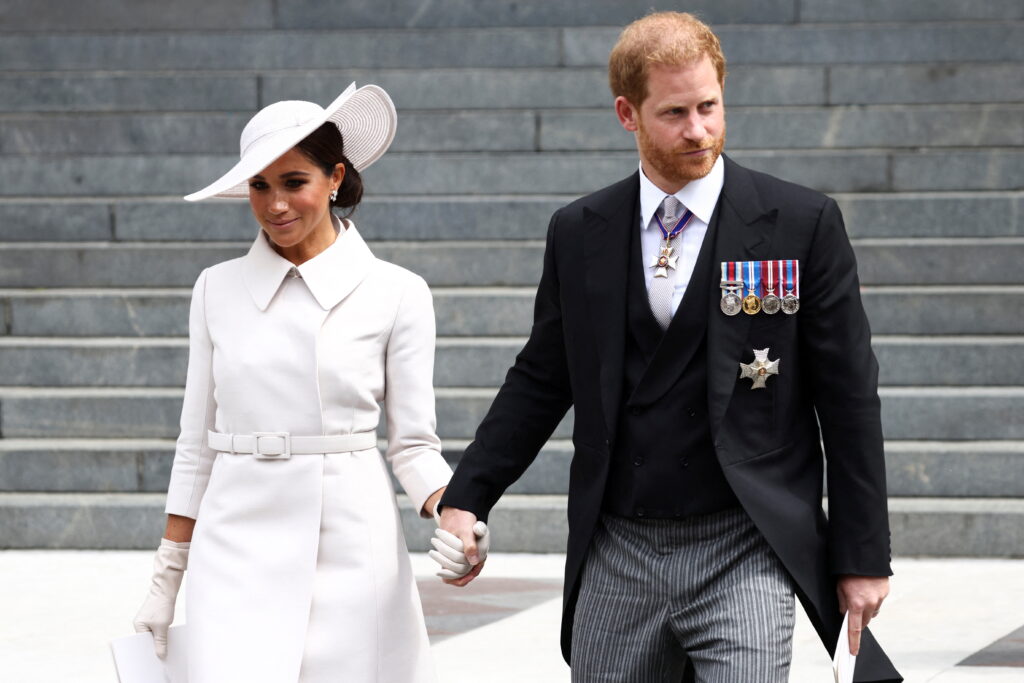 Meghan Markle (L) wearing all-white outfit with matching hat, holding hands with Prince Harry, who is dressed in a black suit with gray striped pants