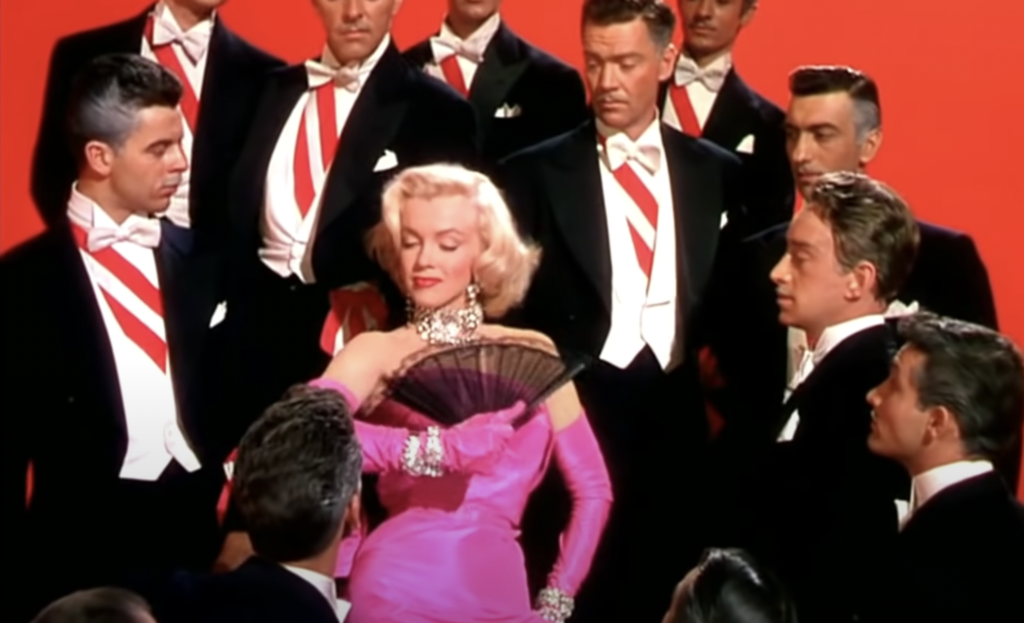 Marilyn Monroe wearing pink dress and holding fan while surrounded by men in white tuxedos and red ties
